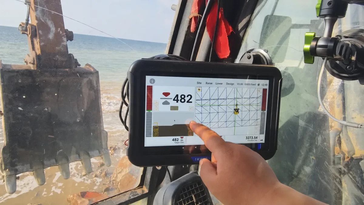 Excavator operator reading GNSS cabin display with mesh design on coastal site